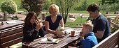 Einen ruhigen Biergarten direkt neben der Minigolfanlage nutzt eine Familie zur Stärkung. Im Hintergund wird der Minigolfplatz von der Sonne bestrahlt.