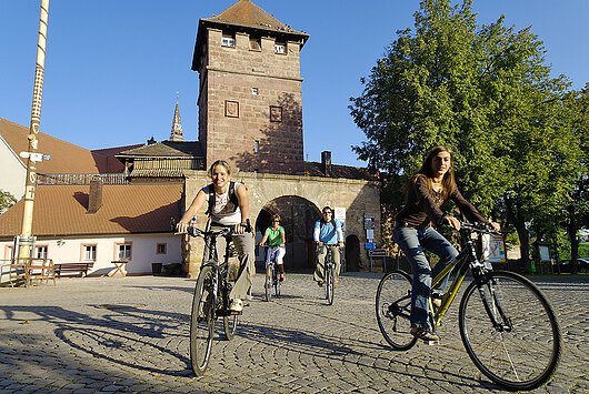 Drei Radfaherer radeln aus der ALtsadt Wolframs-Eschenabch aus dem Stadttor über die gepflasterte Straße vorbei am Kirchweihbaum.