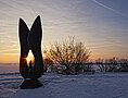 Kunstobjekt bei Sonnenaufgang am schneebedecktem Altmühlsee. Einige Meter dahinter stehen einige Sträucher.