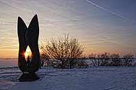 Kunstobjekt bei Sonnenaufgang am schneebedecktem Altmühlsee. Einige Meter dahinter stehen einige Sträucher.