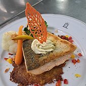 Ein rosa gebratenes Stück Lacks mit einem Blatt und Merrettich dekoriert, liegt auf einem Kartoffelsrösti. Das weiße Teller ist zudem mit Blumenkohl, Karotte und Paprika dekoriert.