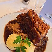 Zwei Kartoffelknödel mit Petersilie garniert liegen auf Bratensoße. Dahinter ein schönes, knuspriges Stück Schäuferle. Ein Sektglas ist zur Hälfte zu sehen.