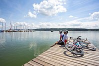 Zwei Rennradler machen Pause am Brombachsee. Sie sitzen dabei auf einem Holzsteg und blicken zu den Segelbooten