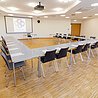 Konferenzraum mit U-förmiger Tischanordnung, Stühlen, Notizblöcken und Gläsern, Projektor und Leinwand an der Wand.
