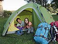 Zwei Personen liegen in einem grünen Zelt, davor Wanderschuhe und zwei große Rucksäcke auf Gras.