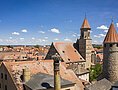 Stadtansicht mit roten Ziegeldächern, Kirchturm und rundem Steinturm unter blauem Himmel mit Wolken.