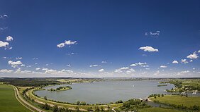 Panoramablick auf einen See mit Uferwegen, Wiesen und blauem Himmel mit vereinzelten Wolken.