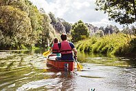 Zwei Personen mit roten Schwimmwesten paddeln in einem roten Kanu auf einem Fluss, umgeben von gr&uuml;ner Natur.