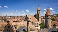 Stadtansicht mit roten Ziegeldächern, Kirchturm und rundem Steinturm unter blauem Himmel mit Wolken.
