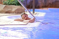 Frau lehnt im Schwimmbad am Beckenrand und stützt den Kopf auf die Arme im Wasser.