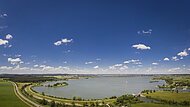 Panoramablick auf einen See mit Uferwegen, Wiesen und blauem Himmel mit vereinzelten Wolken.