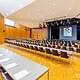 Gro&szlig;er Konferenzraum mit Reihen von Tischen und St&uuml;hlen, B&uuml;hne mit Rednerpult und Leinwand im Hintergrund.