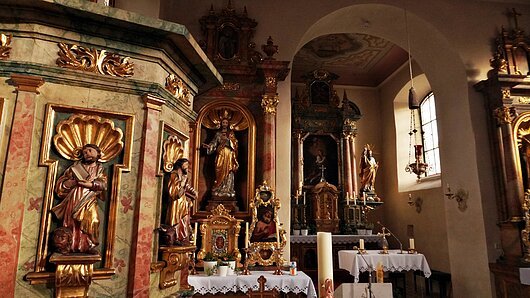 Innenansicht einer Kirche mit Alt&auml;ren, goldverzierten Heiligenfiguren und Kerzen auf Tischen mit wei&szlig;en T&uuml;chern