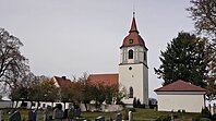 Kirche mit Turm und rotem Dach in einem Friedhof mit Grabsteinen und B&auml;umen bei bew&ouml;lktem Himmel.