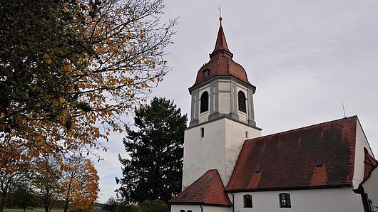 Kirchturm mit rotem Dach neben B&auml;umen mit herbstlichen Bl&auml;ttern unter bew&ouml;lktem Himmel.